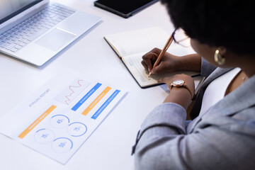 Writing in notepad, businesswoman analyzing annual report at office desk
