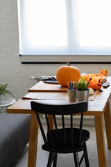 A wooden table with a pumpkin, craft supplies, and Halloween decorations sits in a bright room near a black chair. A cozy, minimalist setting with natural light from large windows.