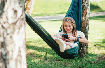 Little girl enjoys sunny day swinging in hammock and browsing internet using cell phone in serene...