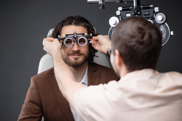 Doctor ophthalmologist checks vision of happy young man in armchair of modern optician clinic for...