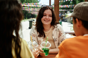 Portrait shot of charming teenage girl sitting at cafe table in park listening with smile to friends while enjoying time together and summer day outside, copy space