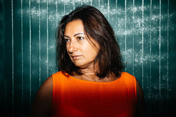 Portrait of woman wearing orange top against green background with shadows, thoughtful expression,...