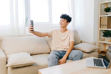 Young man taking a selfie in a bright living room, surrounded by soft furnishings, natural light enhancing the cheerful atmosphere and casual vibe of his home environment