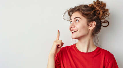 Obraz premium A young woman with her hair in a messy bun smiles brightly, pointing upwards, wearing a red shirt. The image has a light minimalist background