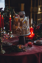Elegant luxury Christmas table setting for family home party with homemade big gingerbread house, holiday celebration. Red tablecloth, candles, chandelier. Romantic candlelit dinner