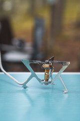 A compact camping stove stands on a blue table in a wooded area, prepared for cooking outdoors during fall