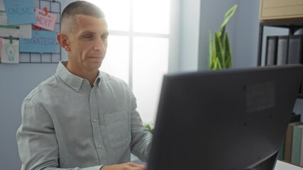 Man working on computer in bright modern office, surrounded by motivational posters, organized workspace, and natural light from large window