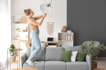 Young woman on sofa screwing light bulb into hanging lamp at home