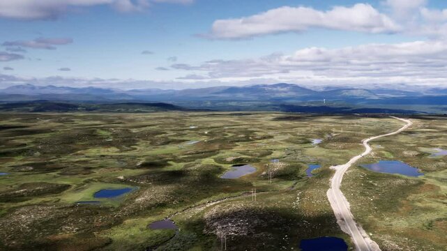 Drone flying sideways as camera zooms out, dirt road winding through the tundra landscape of the Flatruet High Fell plateau. With ponds, heathland and the mountains on the horizon in J&auml;mtland Sweden