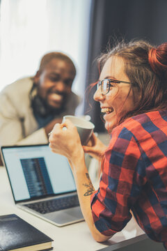 A woman with glasses and a red plaid shirt is smiling while holding a coffee cup. She is sitting next to a laptop computer