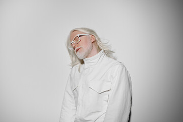 Portrait of a person with long white hair wearing a white jacket against a light gray backdrop,...