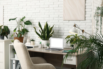 Workplace with blank laptop, lamp and green plants near white brick wall in office