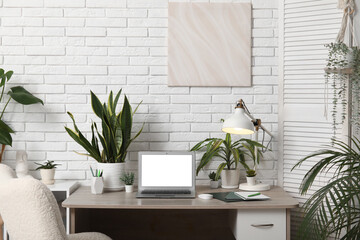 Workplace with blank laptop, lamp and green plants near white brick wall in office