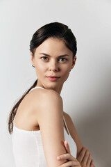 Portrait of a confident young woman with a serene expression, wearing a white tank top, showcasing natural beauty against a light gray background