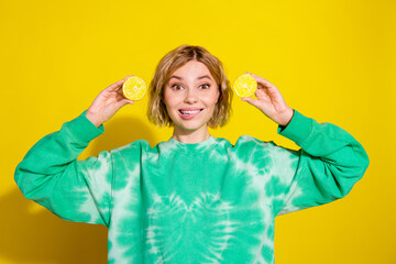 Photo of cheerful pretty lady dressed green sweatshirt showing two lemon slices biting lip empty space isolated yellow color background