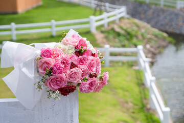 Pink flower bouquet rests on a white fence, with a grassy outdoor setting and water in the background.