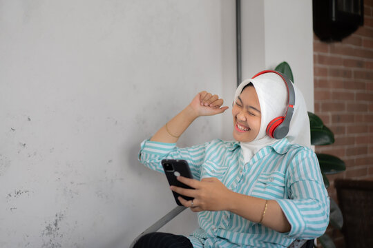 A woman wearing hijab and headphones is smiling while looking at her cell phone. She is happy and enjoying music. copy space is available for advertising words. in casual spaces such as cafes or hango