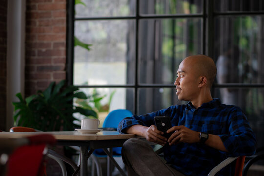 Thoughtful bald man in a plaid shirt relaxing in a modern loft-style cafe, holding a smartphone and looking out the window during a coffee break.