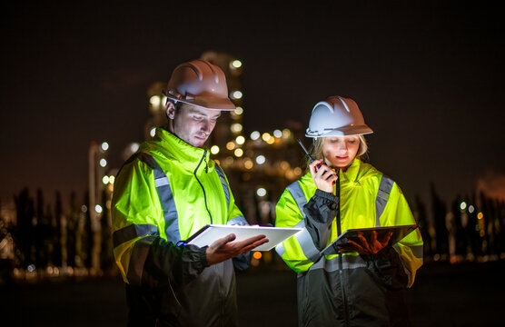 Engineers wearing safety gear, including hard hats examining survey are using tablet collaborating and discussing on screen work plant site use with night lights of oil refinery industry background.
