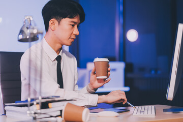 Young pensive asian man working late concentrated and serious in front of computer at night in dark...