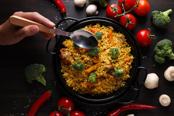 Pilaf in frying pan, vegetables and hand with spoon on dark wooden background, top view