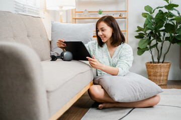 A young woman sitting on the floor, leaning against a sofa, using a tablet in a modern living room with cozy decor and an indoor plant.