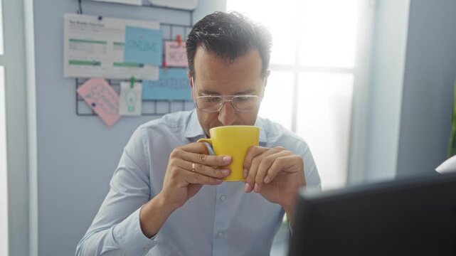 Middle-aged man in office drinking from yellow mug, wearing glasses, sitting at desk with background of pinned notes and documents, indoor workspace with light blue walls