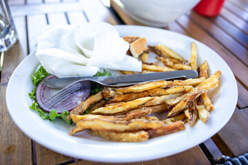 Leftover unfinished plate of french fries and hamburger veggies used utensils and crumbled up napkin sitting on dirty white plate.