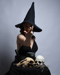 Close up portrait of beautiful  female model wearing black dress, pointy witch hat halloween costume.  Isolated figure on studio background. Casting magic spell at spooky occult  ritual potion table 