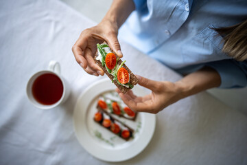 Woman having breakfast, lunch, snacking on delicious rye crisp bread sandwich with cream cheese, cherry tomatoes, arugula, basil, parsley. Calm girl eating nutritious nourishing food at morning.