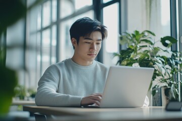 Fototapeta premium Young Asian businessman working on laptop at minimalist desk in modern office
