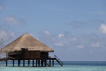 A wooden overwater bungalow with a thatched roof, situated above clear turquoise waters. The sky is partly cloudy, creating a serene tropical atmosphere.