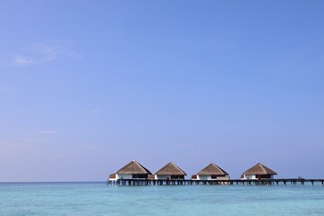 A serene view of overwater bungalows on stilts in a clear turquoise sea under a bright blue sky. The scene conveys tranquility and tropical paradise.