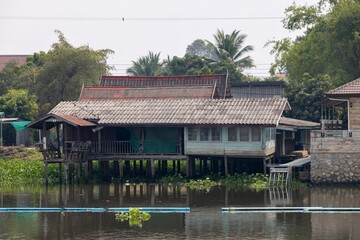 A traditional wooden house on stilts by a river, surrounded by lush greenery. The house features a tiled roof and open balcony, reflecting local architecture.