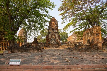A serene ancient temple complex with crumbling brick structures, surrounded by lush green trees and fallen leaves. Statues and remnants of historical architecture are visible, creating a tranquil atmo