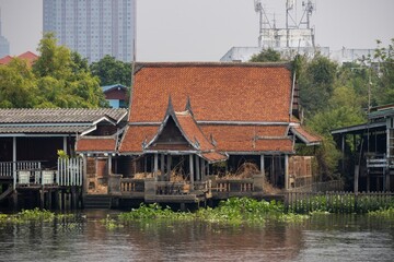 A traditional Thai wooden house on stilts by a river, featuring a distinctive red-tiled roof and surrounded by lush greenery and water lilies. The scene captures the serene beauty of Thai architecture