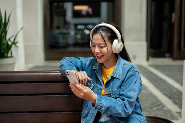 A woman is listening to music and reading a chat on her phone while sitting on a bench in the city.