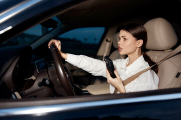 Woman with cell phone sitting in car, hands on steering wheel, focusing on phone