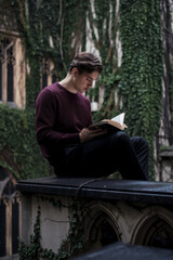 Fototapeta premium Young man sitting on a ledge reading a book in a old university with ivy-covered walls. Concept of education and quiet contemplation.