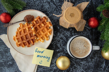 flat lay with a good morning note, a jar of homemade jam, a cup of coffee and homemade Viennese waffles on a black textured background next to Christmas balls and Christmas tree branches. Xmas