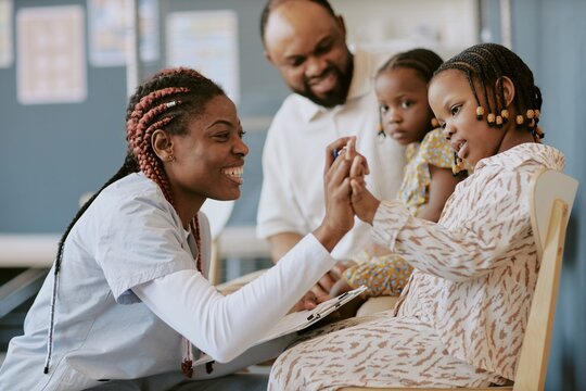 African American nurse engaging with young child using educational toy at medical facility while another child and adult observe interaction attentively
