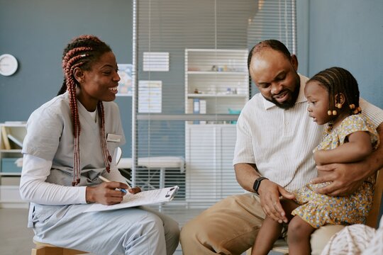 Nurse wearing scrubs speaking with father holding young child in modern clinic setting Perfect lighting emphasizes friendly interaction and welcoming environment