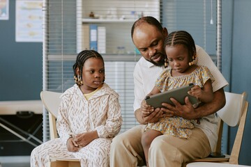 Biracial father spending quality time with daughters teaching them using tablet in living room creating a bonding moment while sitting together on chairs