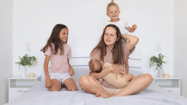 Exhausted mother sitting on bed with hand on head feeling migraine while caring for her infant son and toddler daughters woman grimacing in pain suffering headache kids pulling her hair