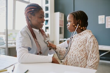 Senior nurse with braids examining young child patient using stethoscope in hospital room smiling...