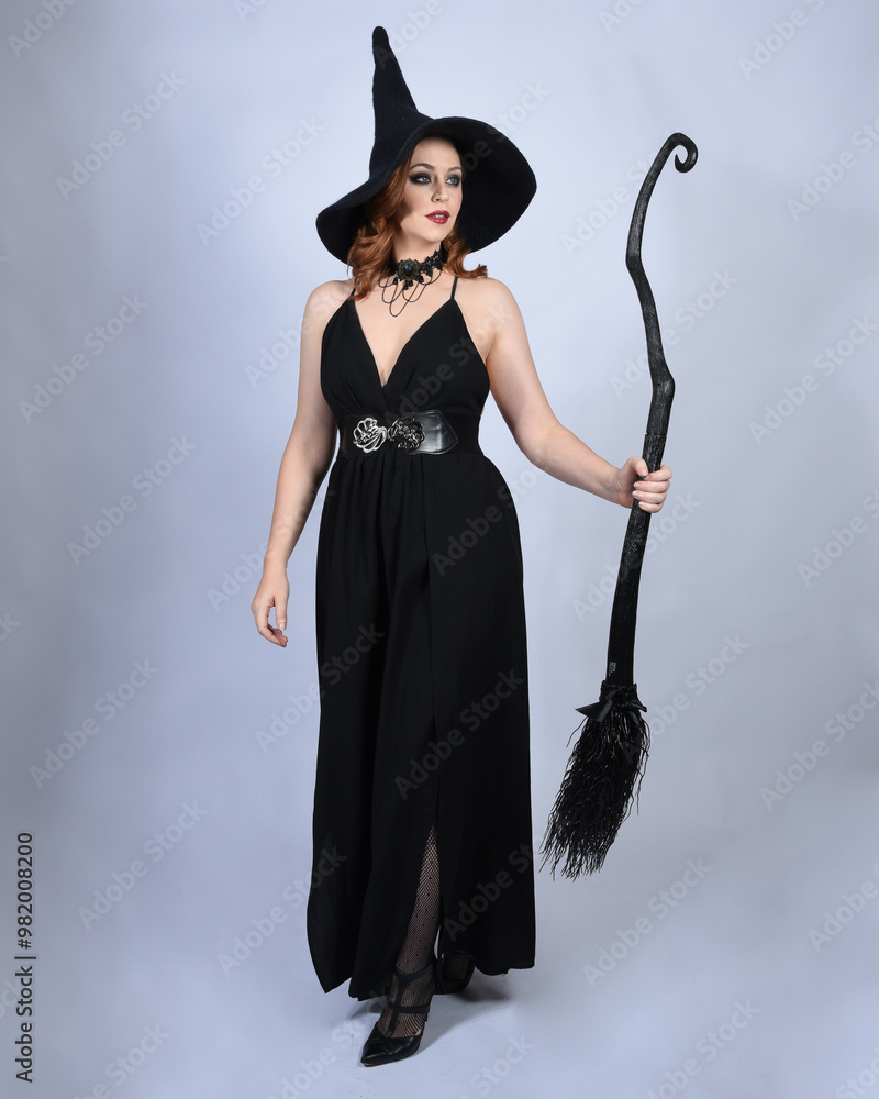 Canvas Prints portrait of beautiful female model wearing glamorous black witch dress, pointy hat halloween costume. walking pose holding broomstick, isolated figure on studio background