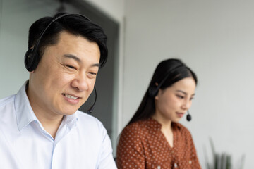 Asian office workers wearing headsets focused on telecommunication tasks. Male and female professionals collaborating in modern office setting, using phone for customer service or technical support.