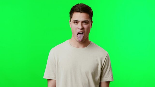 A young man stands against a vibrant green backdrop, playfully sticking his tongue out while making a funny expression, capturing a lighthearted moment of humor.