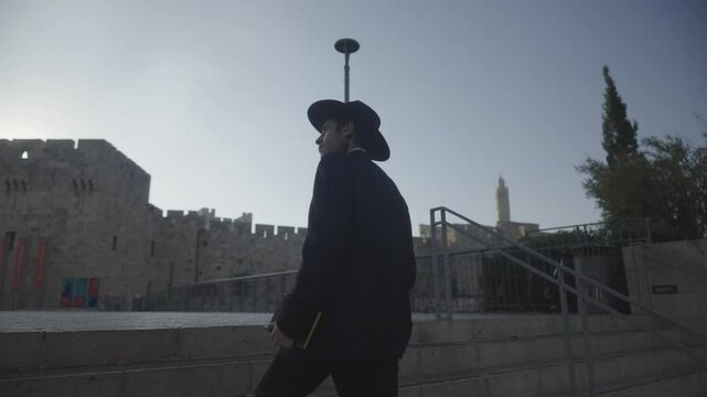 Jerusalem, Israel, an orthodox Jewish man climbs stairs in front of old city and the tower of david walls wearing a black suit and hat and holding a prayer book on a sunny morning