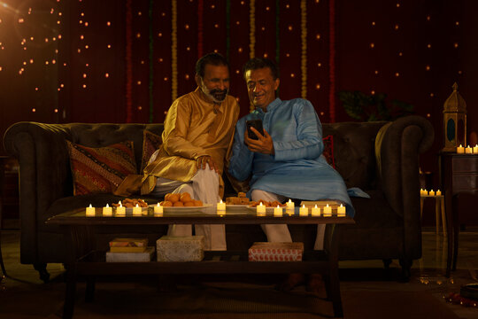 Senior men in traditional outfit sitting together on sofa and using mobile phone while celebrating Diwali festival at home